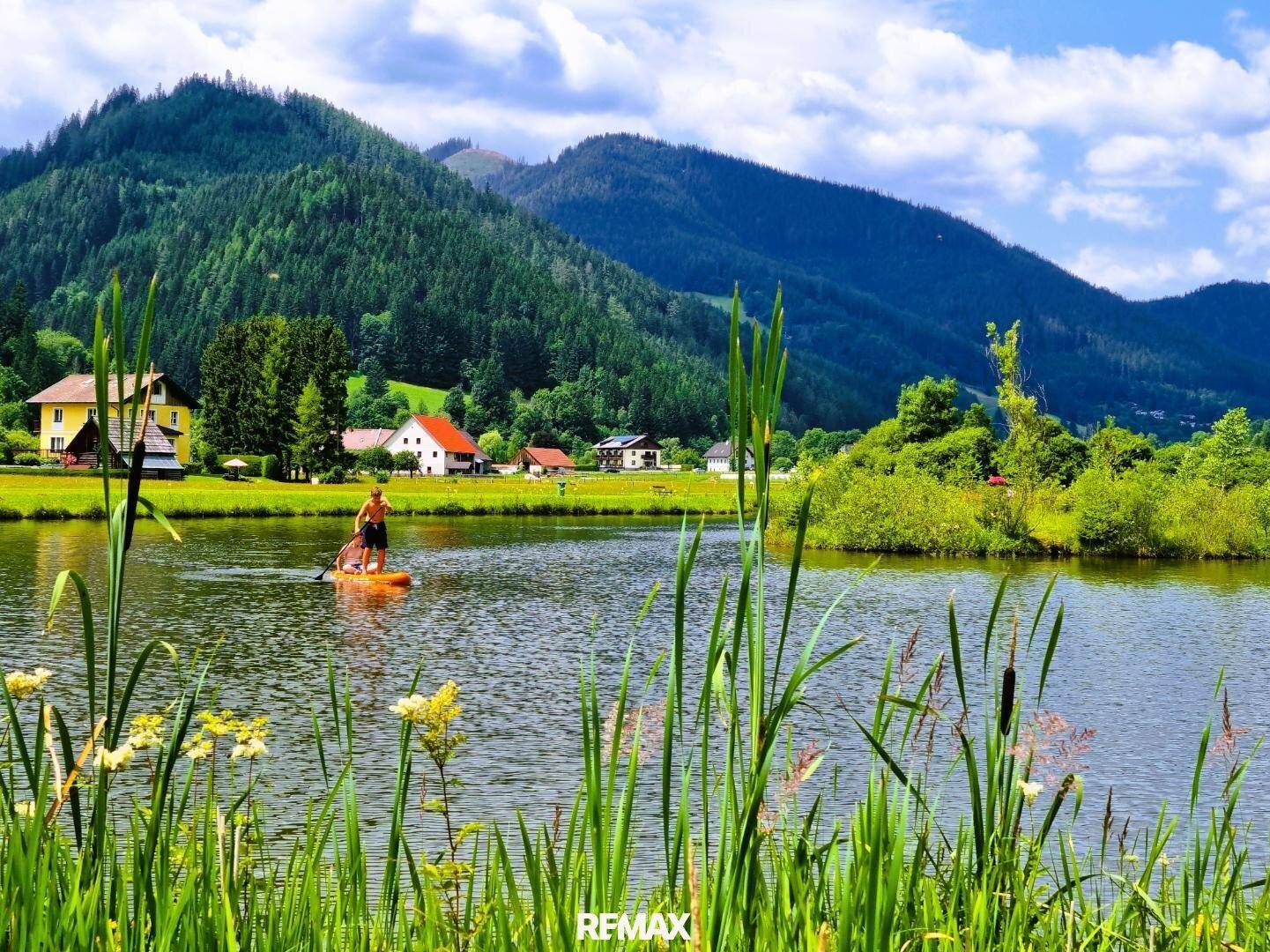 Person beim Stand-Up-Paddling auf einem ruhigen See, umgeben von grünen Hügeln und Häusern im Hintergrund.