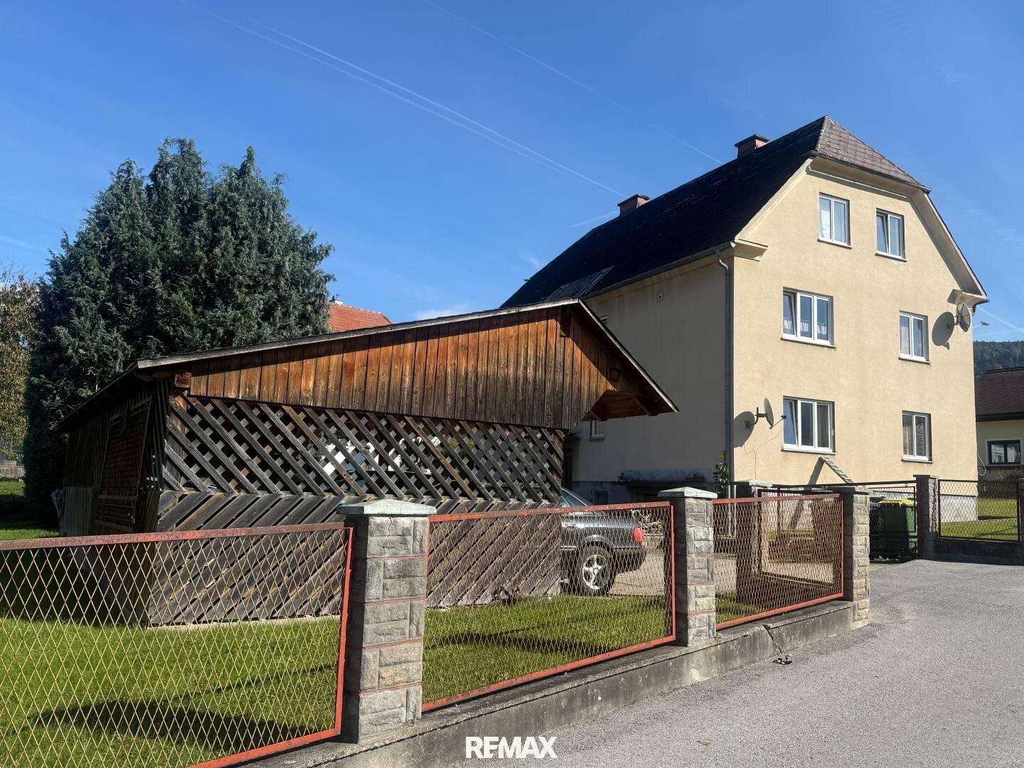 Haus mit Holzcarport und umzäuntem Gartenbereich, umgeben von Grün.