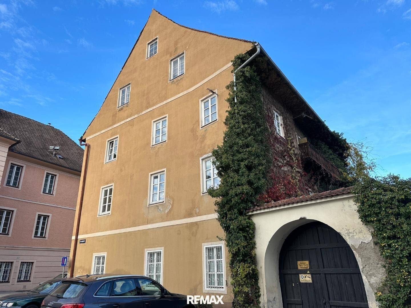 Historisches Gebäude mit ockerfarbener Fassade, grüner Bepflanzung und einem großen Holztor an einem sonnigen Tag.