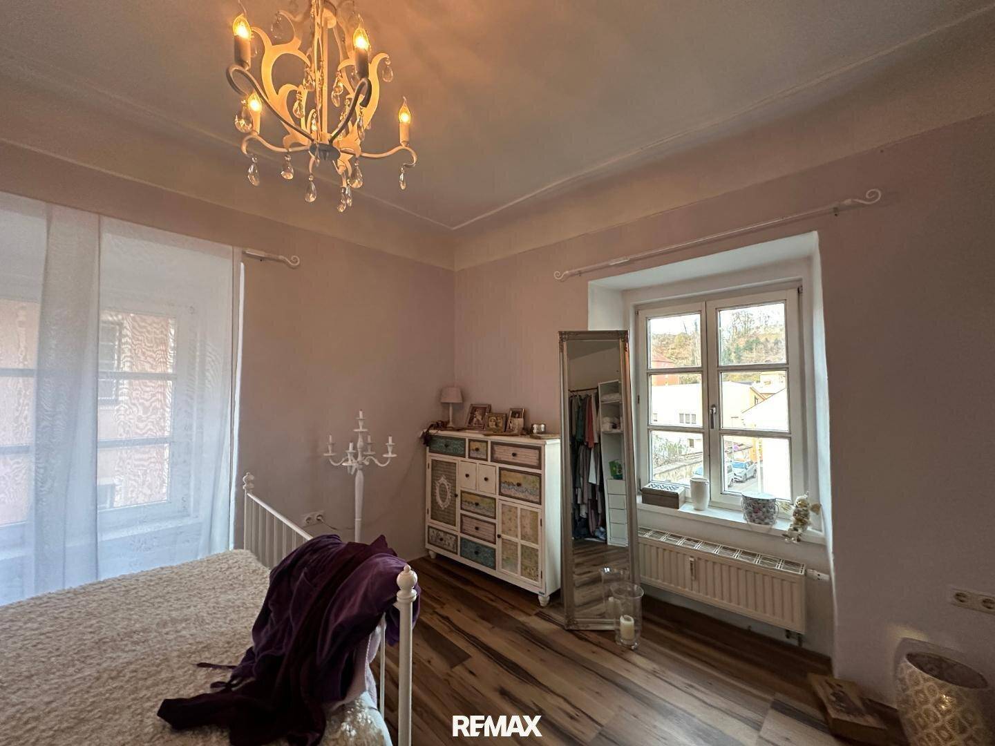 Helles Schlafzimmer mit Holzboden, großem Fenster und einem dekorativen Kronleuchter.