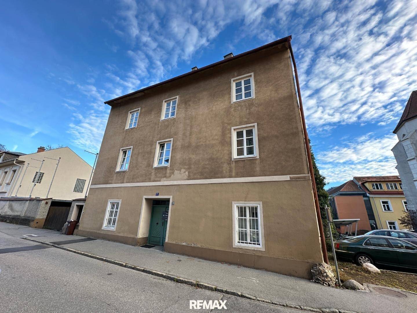 Klassisches Mehrfamilienhaus mit Putzfassade und weißen Sprossenfenstern an einer leicht ansteigenden Straße.
