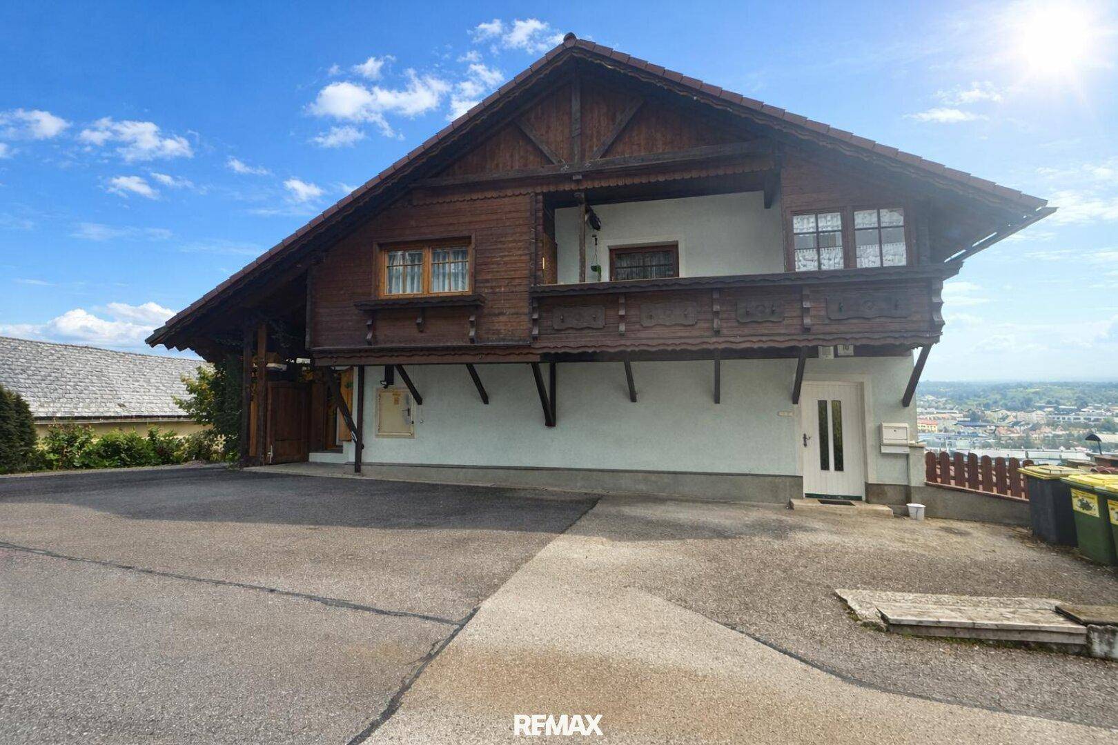 Traditionelles Haus mit Holzbalkon und großem Vorplatz unter strahlend blauem Himmel.