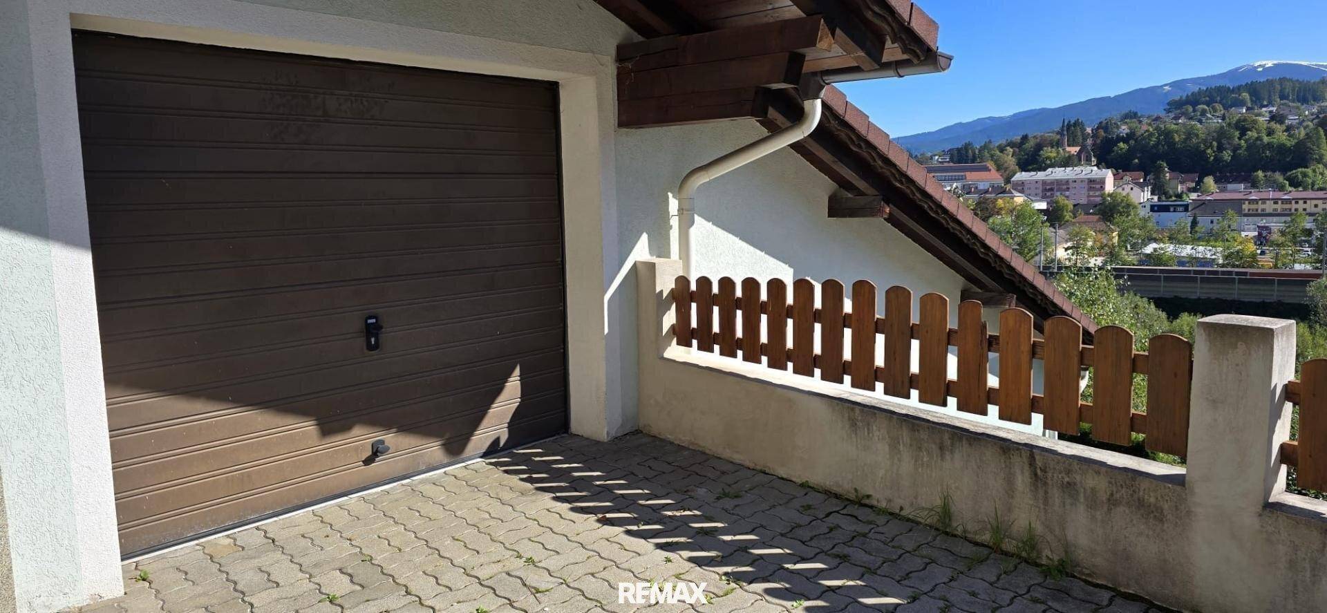 Einzelgarage mit braunem Tor und gepflasterter Zufahrt vor bergiger Kulisse.