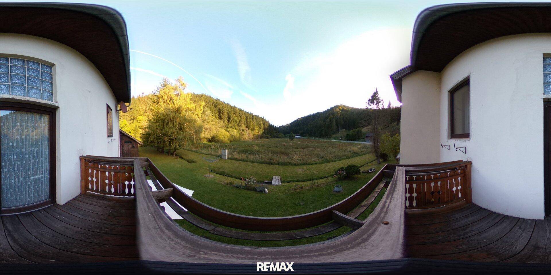 Panoramablick von einem Holzbalcon auf eine weite grüne Landschaft mit Bergen und Wäldern im Hintergrund.