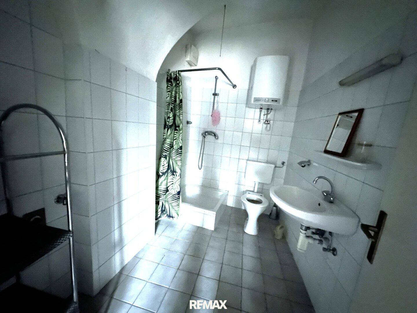 Badezimmer mit weißen Fliesen, Dusche, Waschbecken und Toilette unter einer gewölbten Decke.