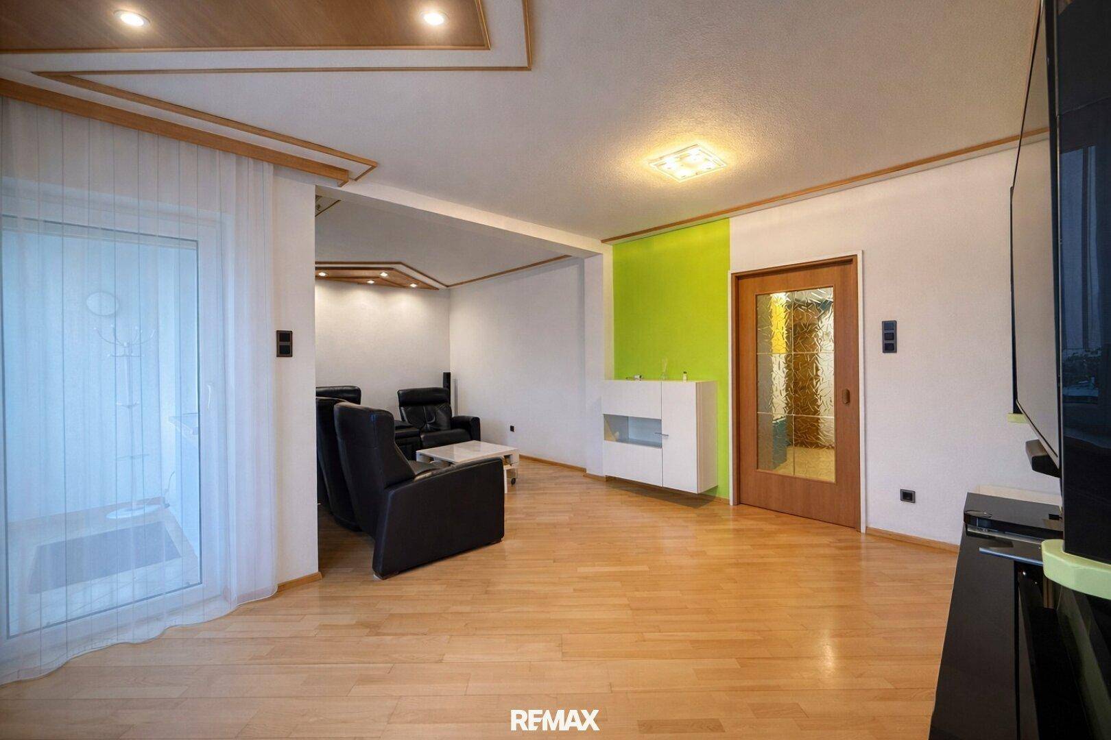 Geräumiges Wohnzimmer mit hellem Holzboden, grüner Akzentwand und Zugang nach draußen.