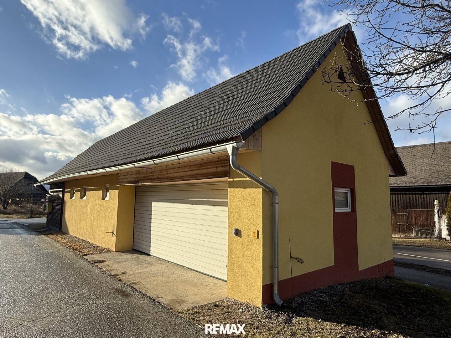 Gelbes Nebengebäude mit Garagentor und Satteldach unter einem bewölkten Himmel.