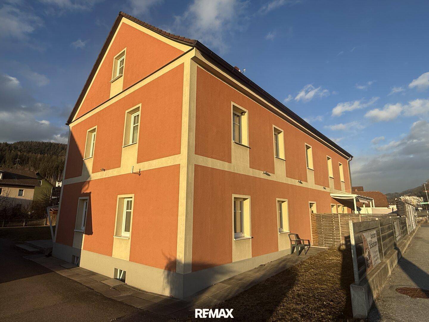 Seitenansicht des Gebäudes mit oranger Fassade und weißen Fensterrahmen unter blauem Himmel.
