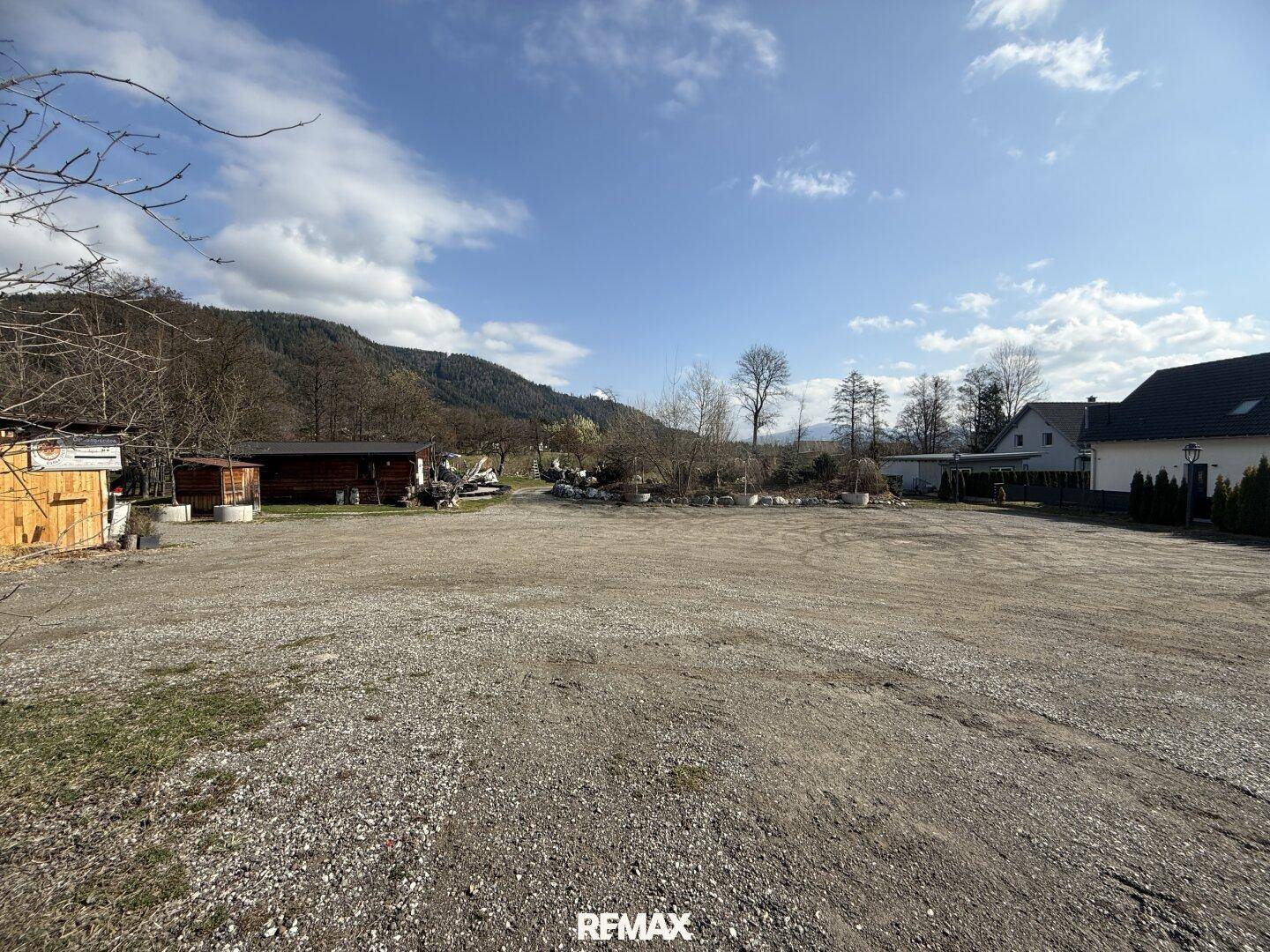 Weitläufiger Schotterparkplatz mit Bergen im Hintergrund und einigen Gebäuden am Rand.