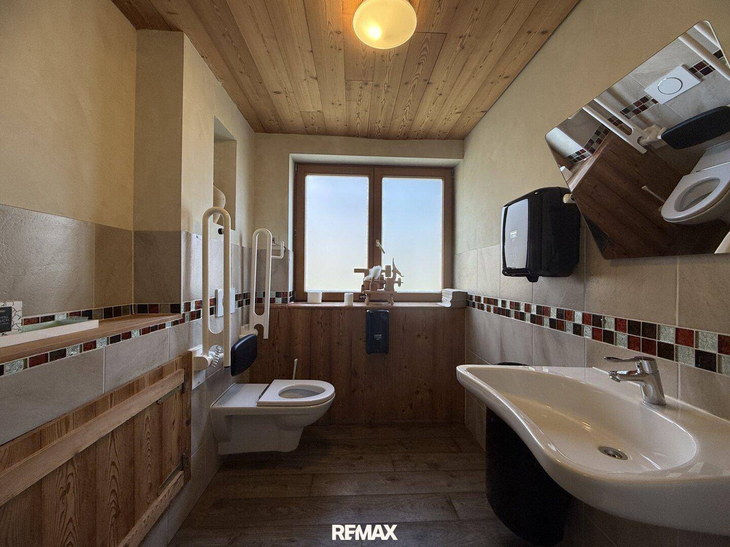 Barrierefreies Badezimmer mit WC, Waschbecken, Haltegriffen und einem Fenster mit Holzakzenten.
