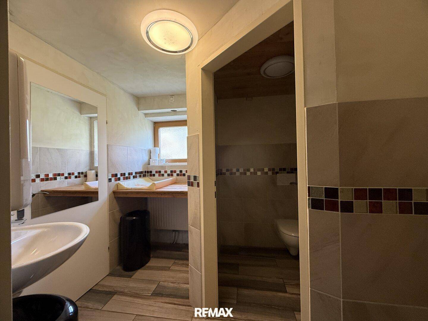 Öffentliche Toilette mit Waschbecken, Spiegel und WC, ausgestattet mit Fliesen und einer Holzdecke.