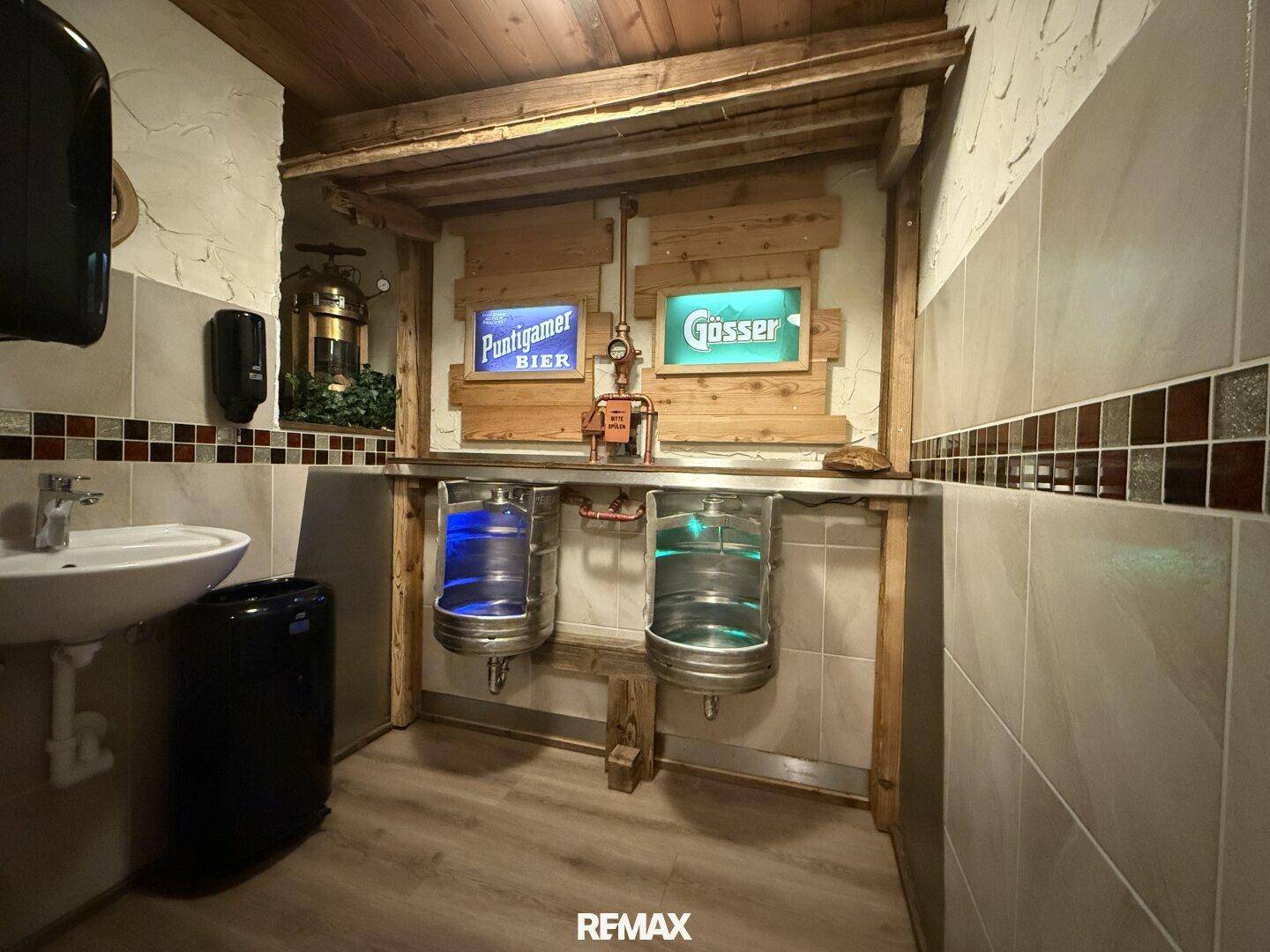 Originelle Herrentoilette mit Bierfass-Urinalen, Waschbecken und gefliesten Wänden in rustikalem Stil.