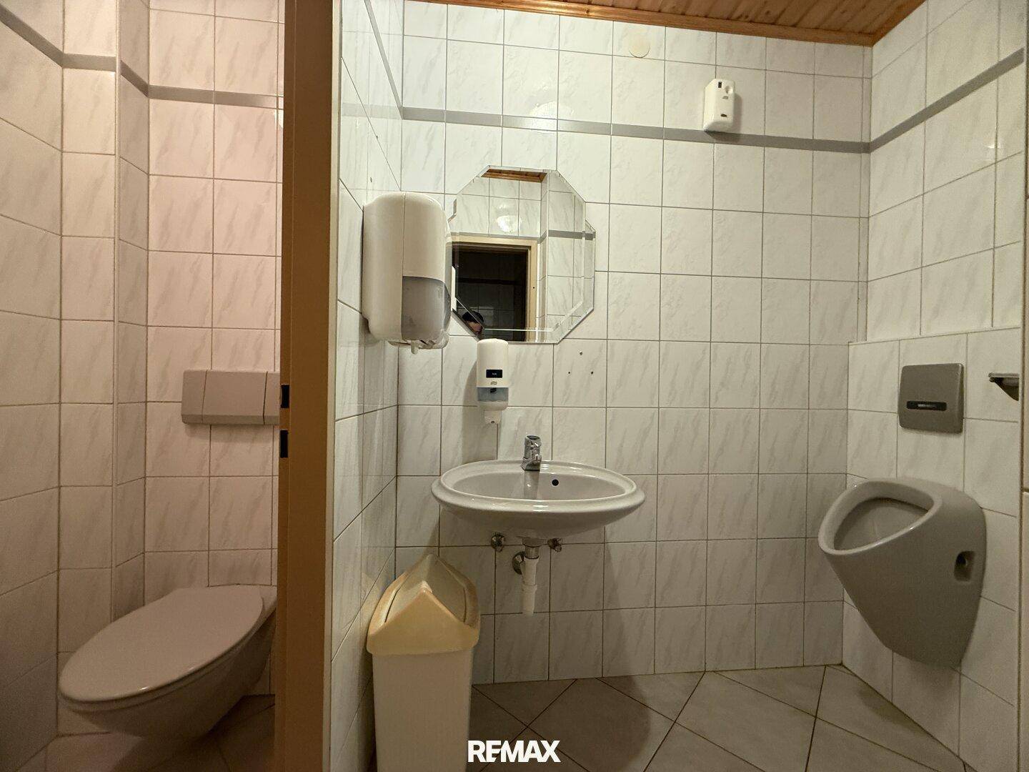 Öffentliche Toilette mit WC, Urinal und Waschbecken, geflieste Wände und eine Holzdecke.