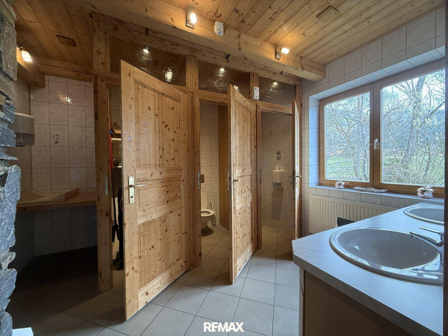 Öffentliche Toiletten mit mehreren Kabinen, Waschbecken und einem Fenster, alles in Holz und Fliesen.