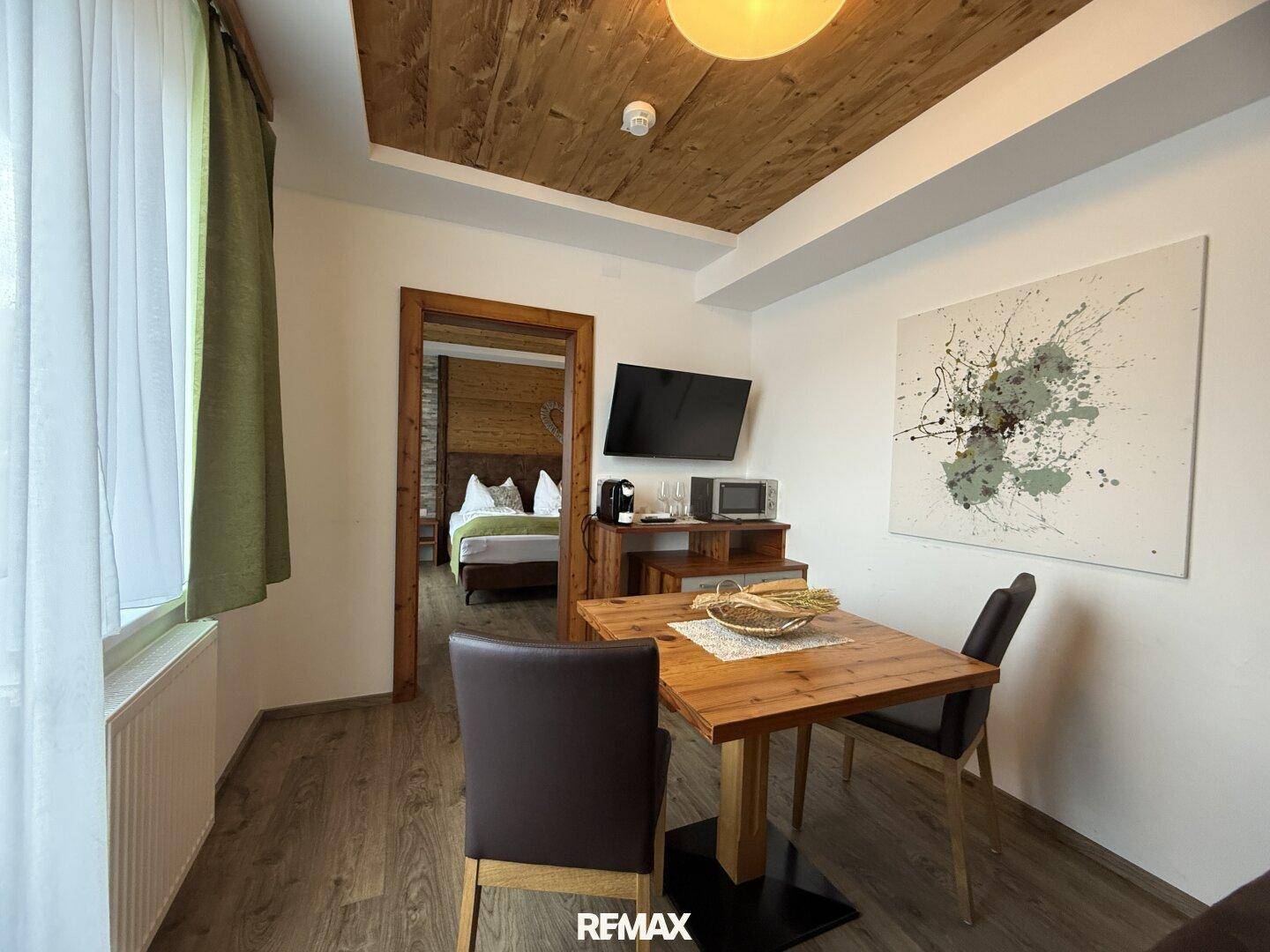 Essbereich mit Holztisch und Blick durch eine offene Tür in ein angrenzendes Schlafzimmer.