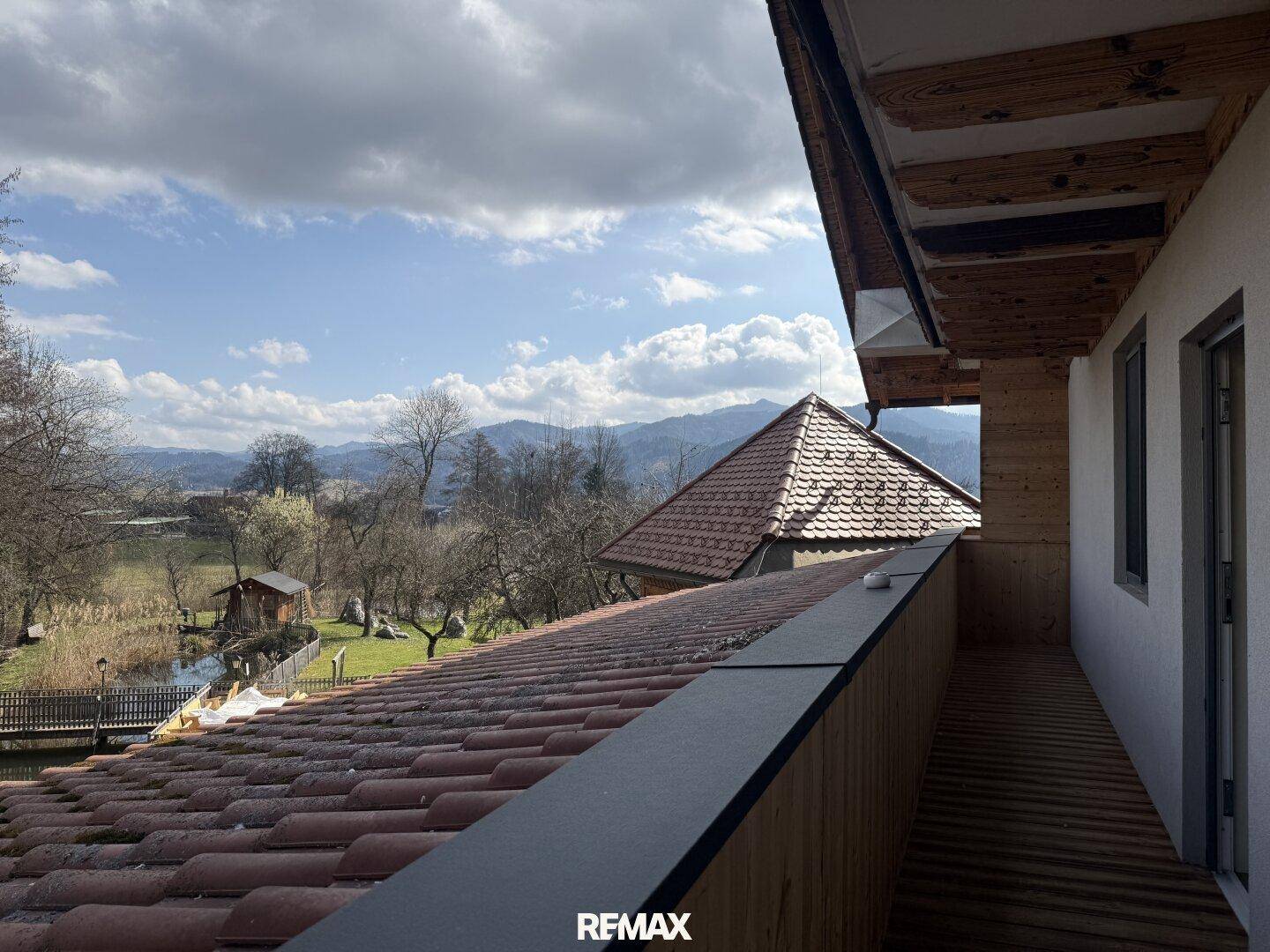 Balkon mit weitem Blick über ein Ziegeldach auf einen See, Bäume und die dahinterliegenden Berge.