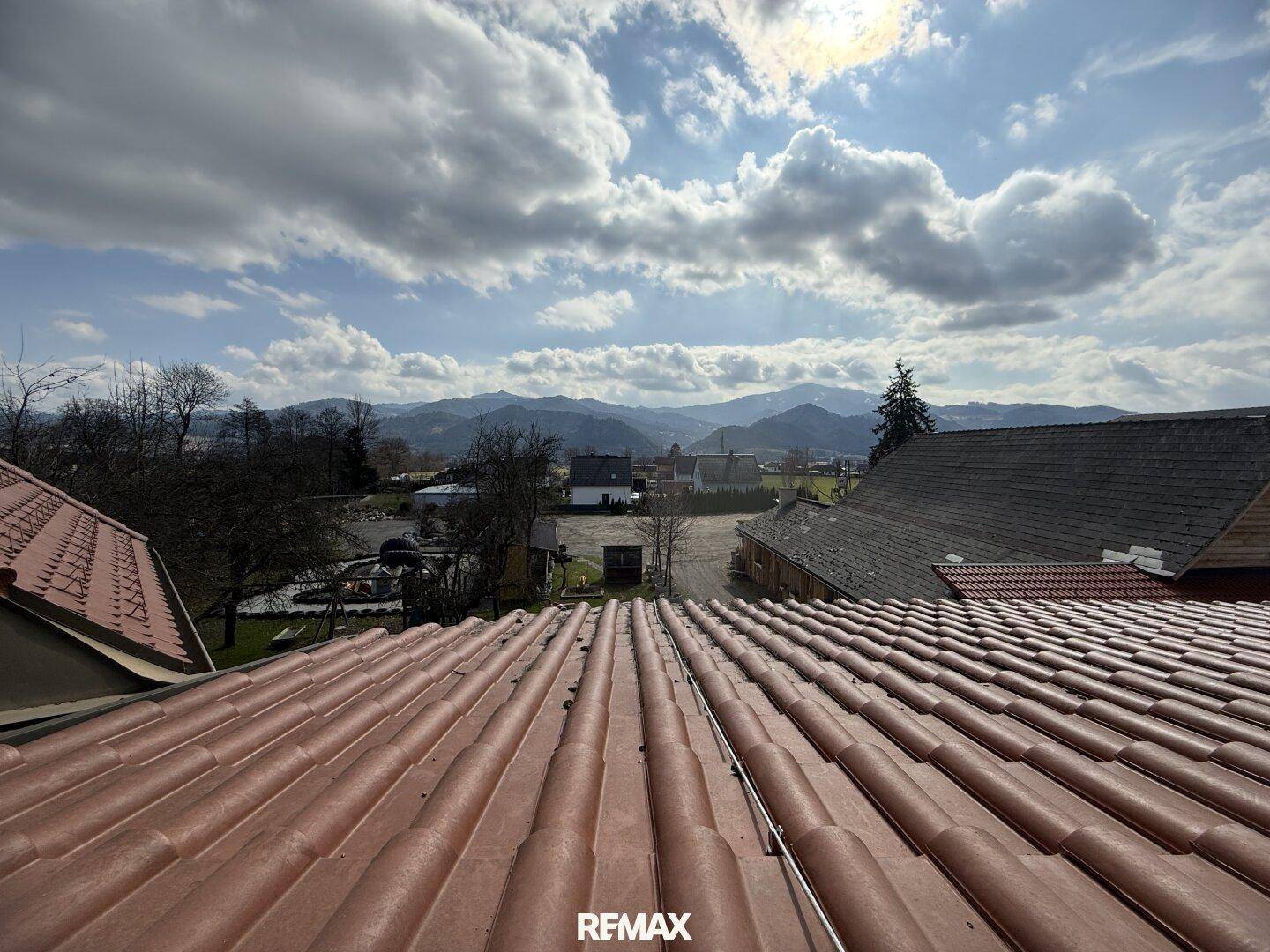 Panoramablick über mehrere Ziegeldächer und eine weite Berglandschaft unter bewölktem Himmel.
