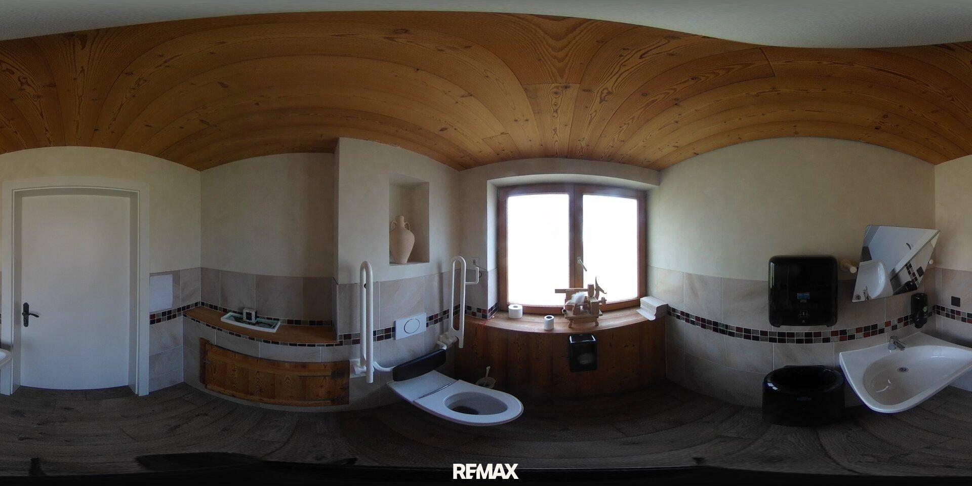 Badezimmer mit Holzbalkendecke, WC, Waschtisch und Haltegriffen für Barrierefreiheit.