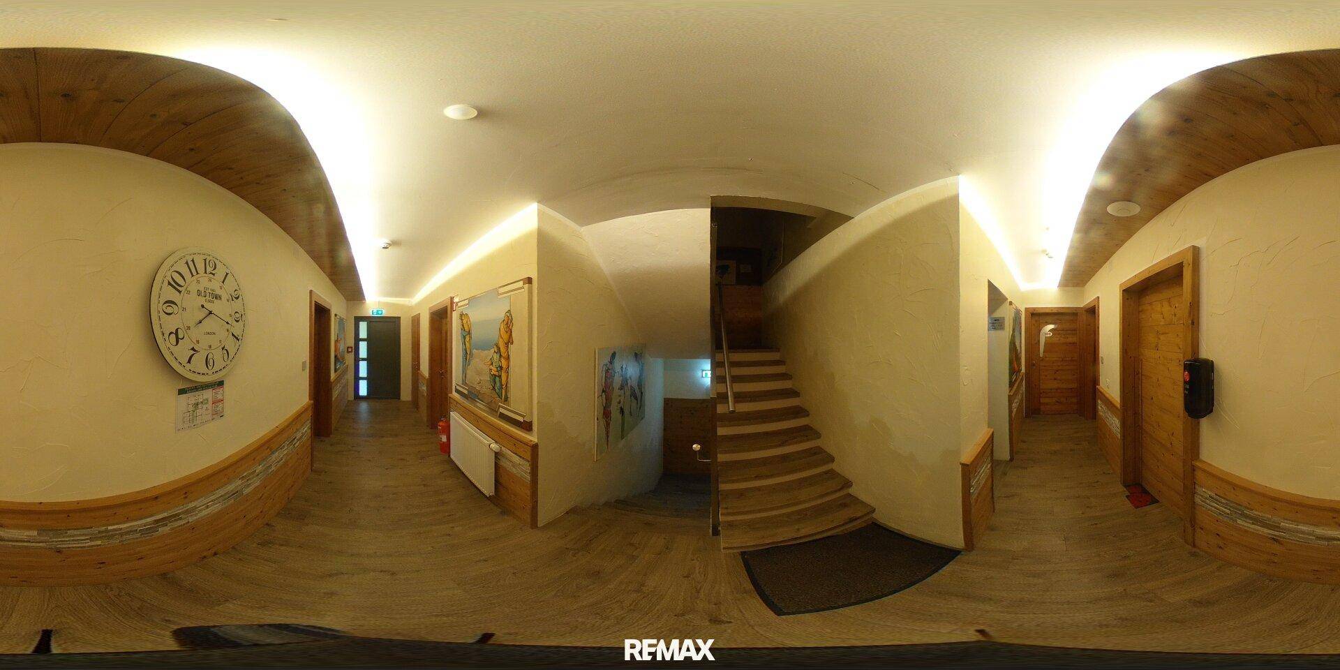 Panoramablick in einen Flur mit großer Wanduhr, Holzvertäfelung und einer einladenden Treppe.