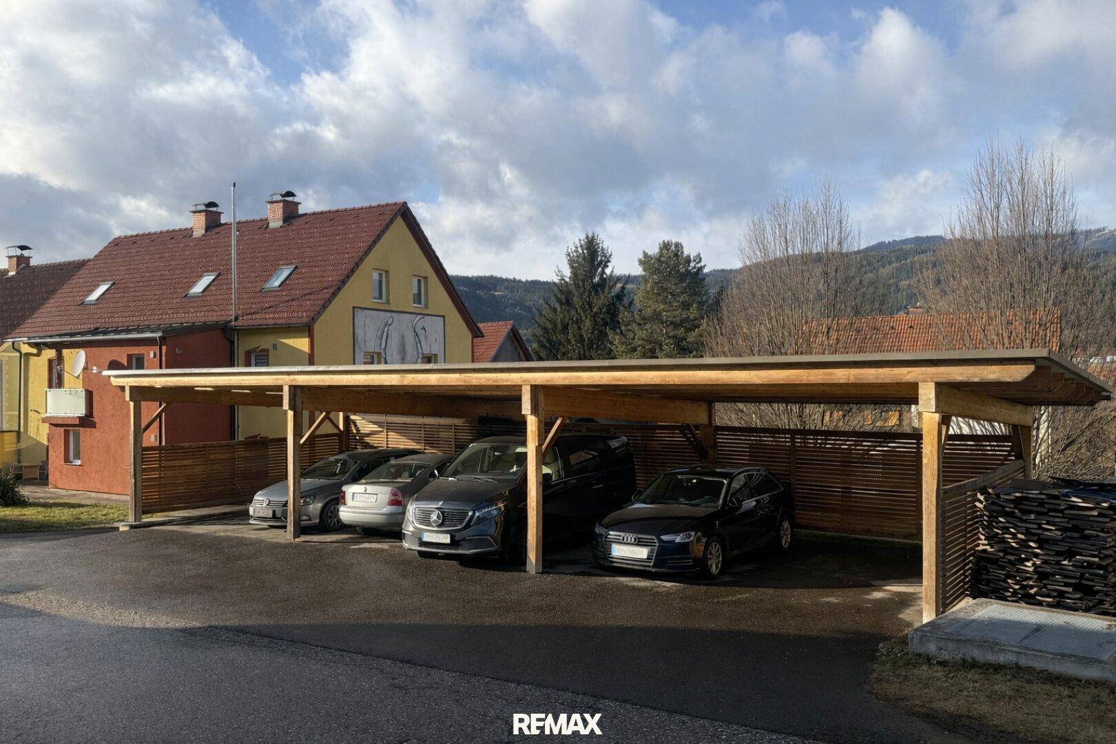 Großer Holzcarport mit mehreren geparkten Autos vor einem Wohnhaus in ländlicher Umgebung.