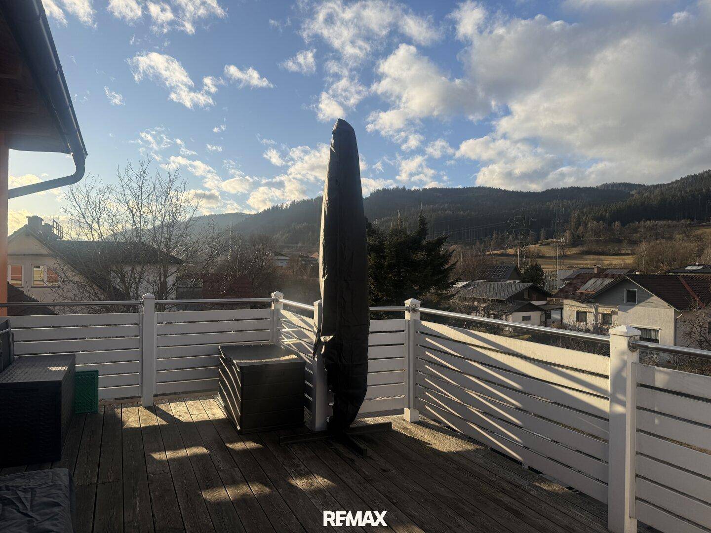 Geräumige Holzterrasse mit weißem Geländer, großem Sonnenschirm und weitem Blick auf die Berge.