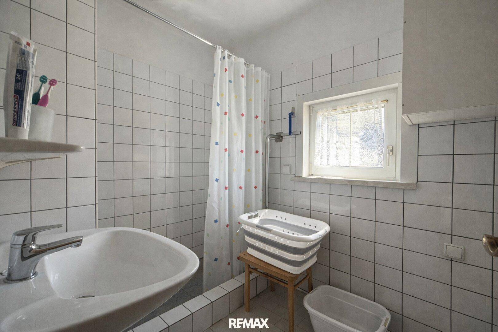Helles Badezimmer mit Duschbereich, Waschbecken und Fenster, komplett gefliest.