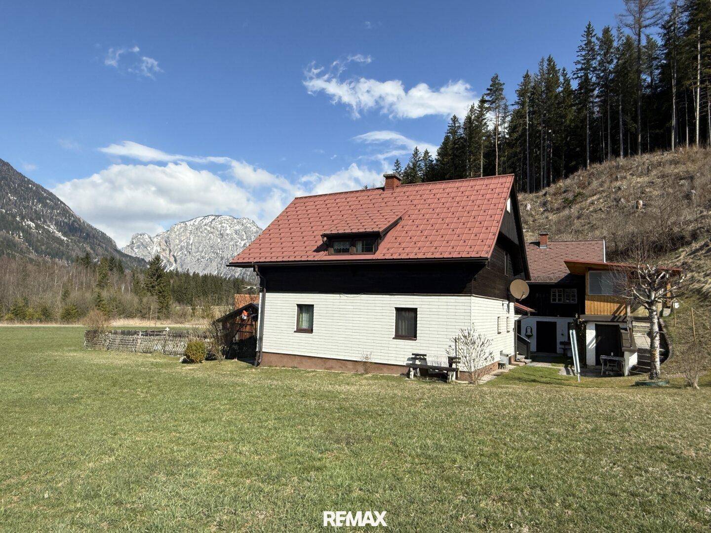 Rückansicht des Hauses mit rotem Ziegeldach und weißer Fassade, umgeben von grünen Wiesen und Bergen.