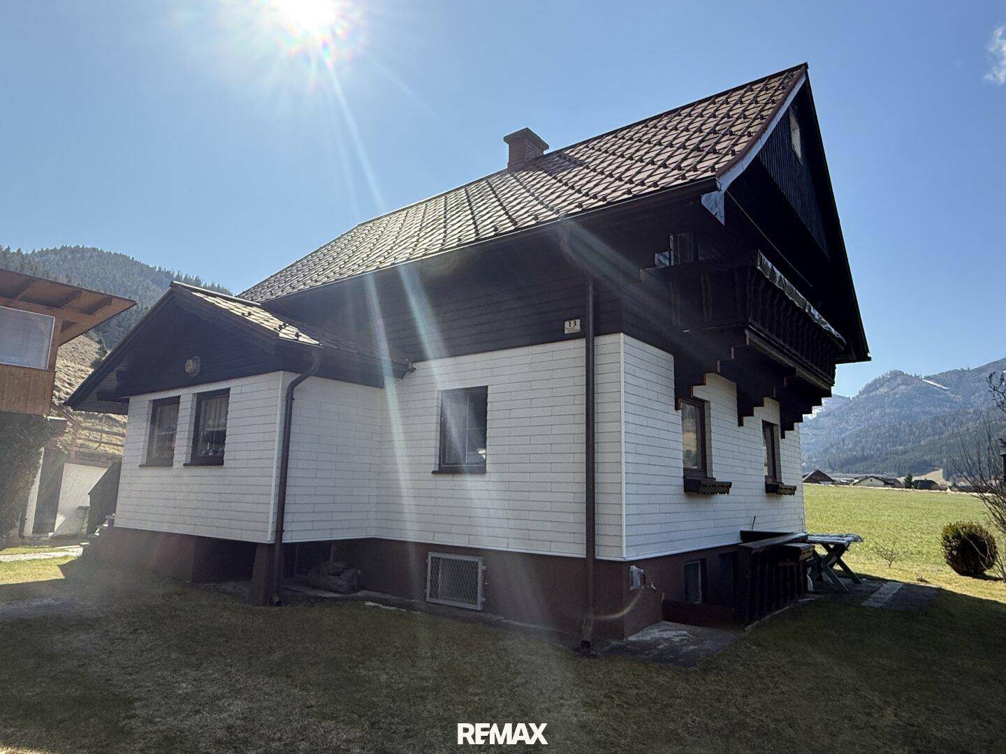 Seitenansicht eines traditionellen Hauses mit Holzbalkon und Ziegeldach in sonniger Berglandschaft.