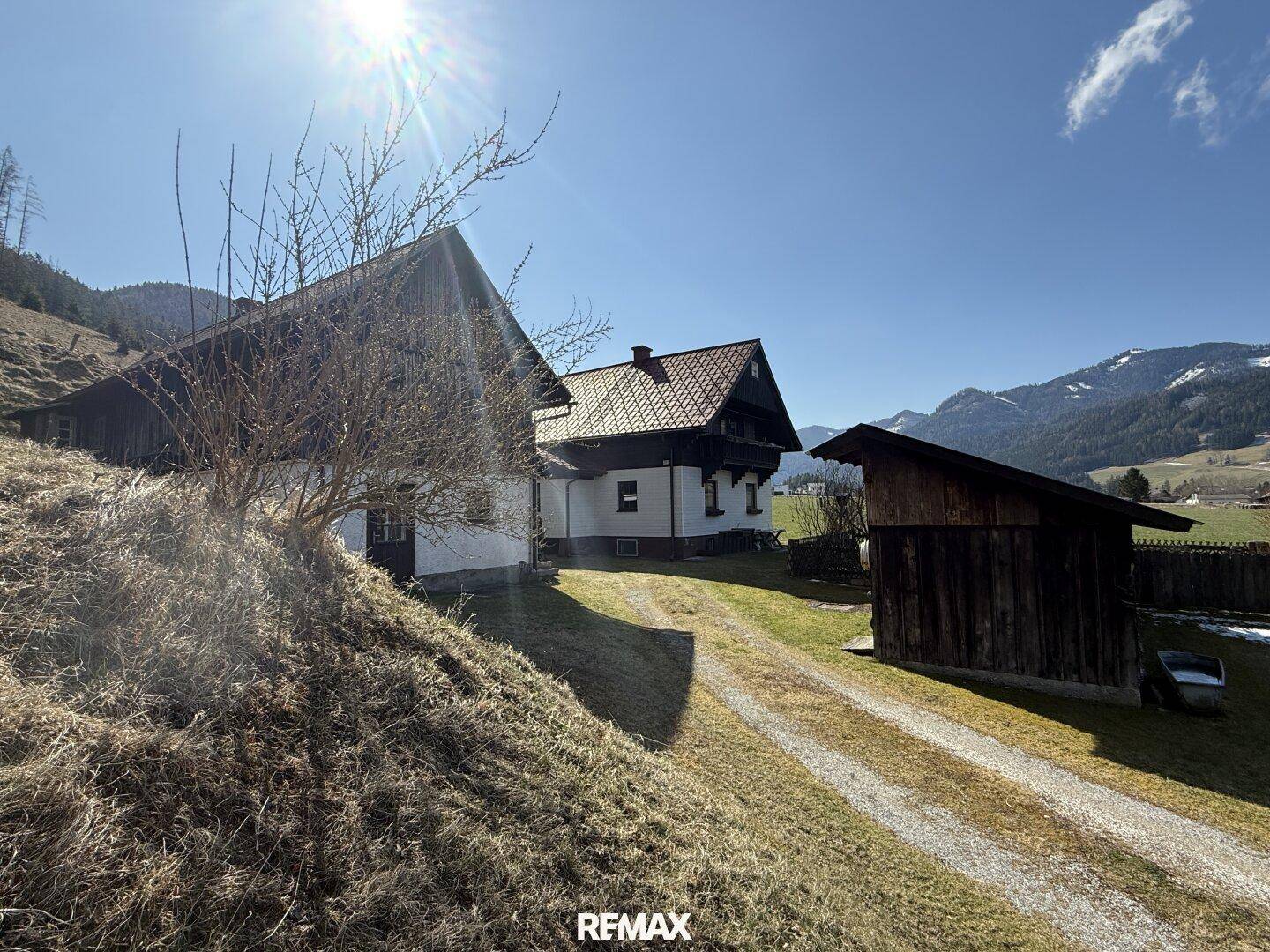 Anwesen mit Haupthaus und Nebengebäude, umgeben von Wiesen und Bergen unter blauem Himmel.
