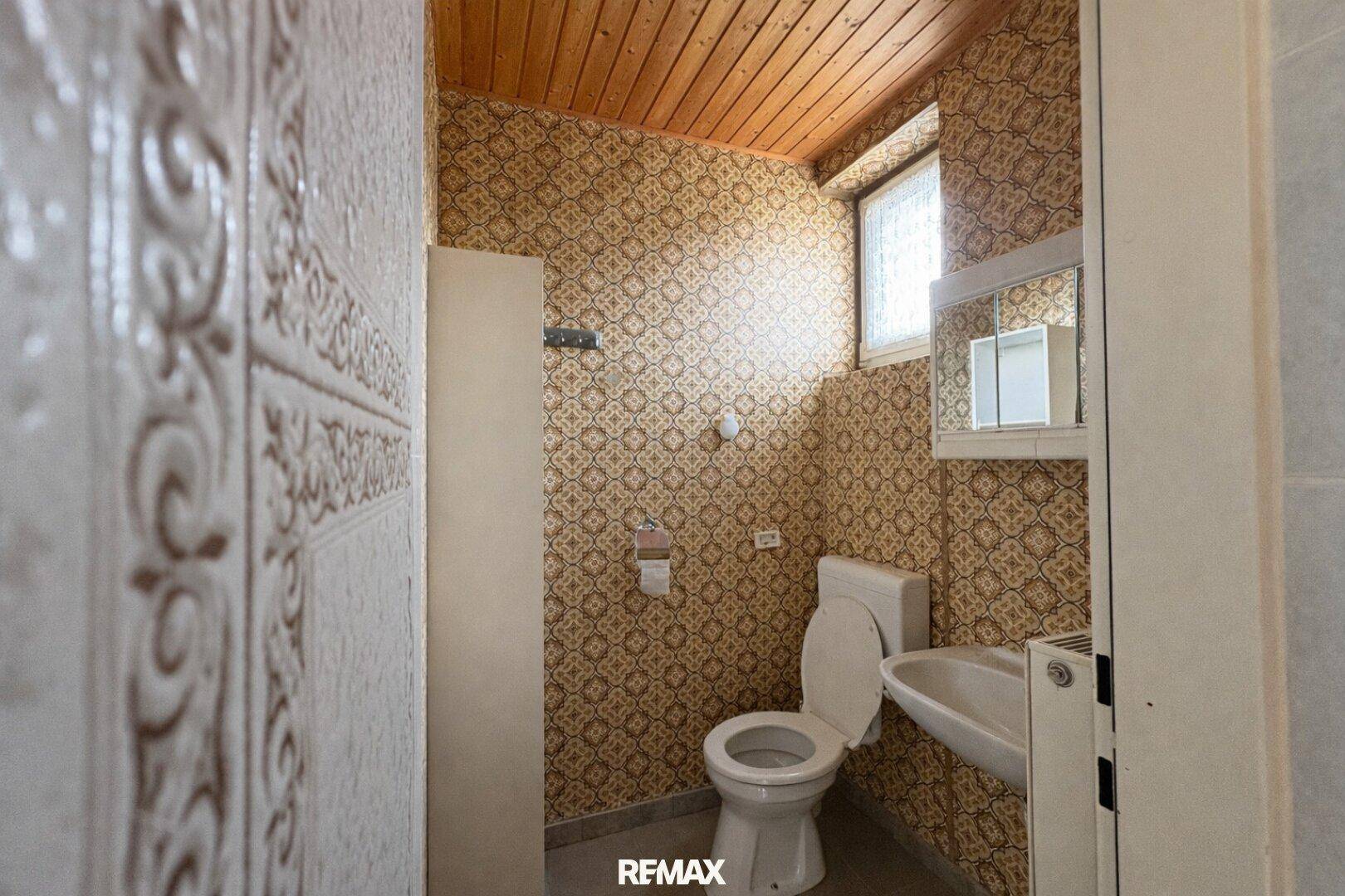 Kleines WC mit gemusterten Fliesen, Toilette, Waschbecken und Fenster.