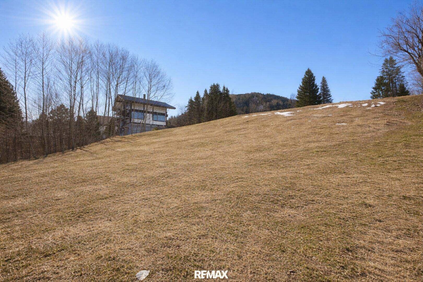 Sonniger Hang mit Blick auf ein modernes Haus, umgeben von Bäumen und ländlicher Landschaft.