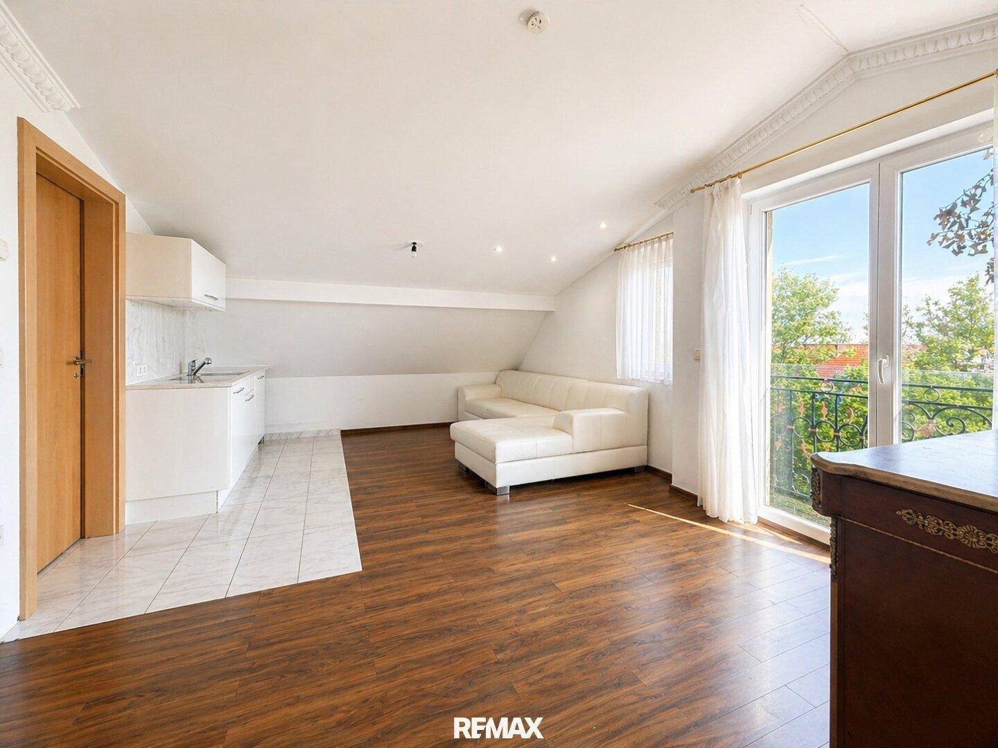 Geräumiger Wohnbereich mit Holzboden, kleiner Küchenzeile und Zugang zu einem französischen Balkon.
