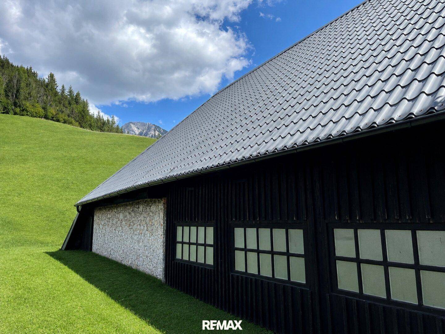 Seitenansicht eines Gebäudes mit dunkler Holzfassade und Ziegeldach vor grüner Hügellandschaft.