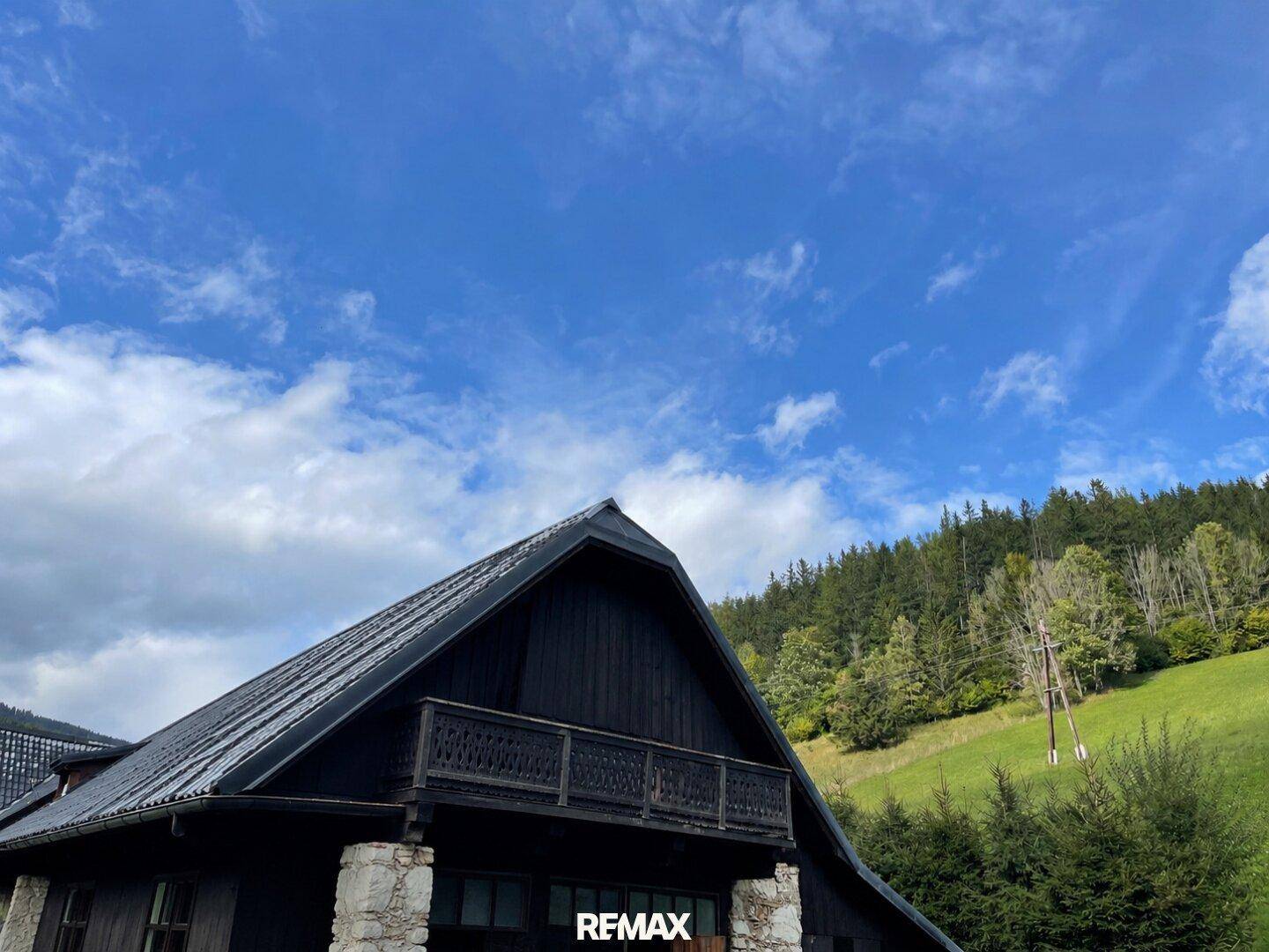 Detailansicht des rustikalen Holzhauses mit dunklem Dach und Holzbalkon vor bewaldetem Hügel.