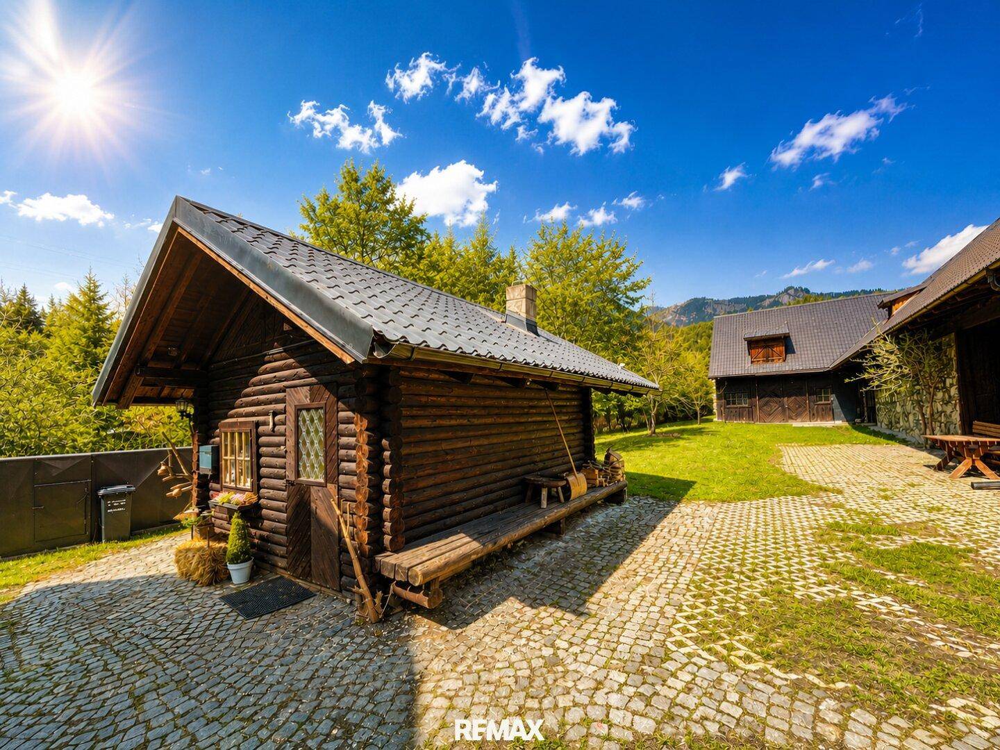 Rustikale Blockhütte mit Holzbänken und gepflastertem Vorplatz in sonniger, grüner Umgebung.
