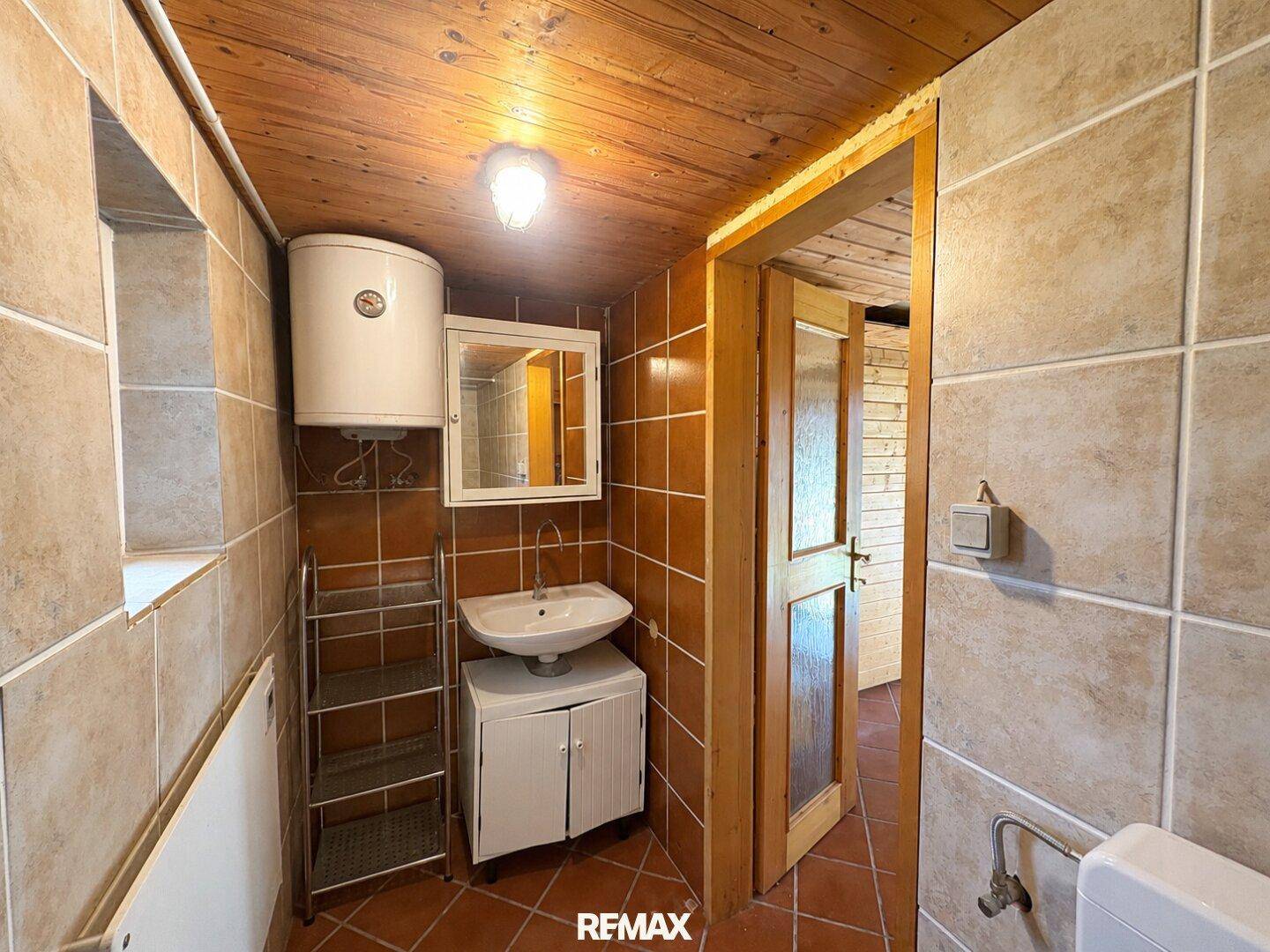 Kleines Badezimmer mit beigefarbenen Fliesen, Waschbecken und Holzdecke, funktional eingerichtet.
