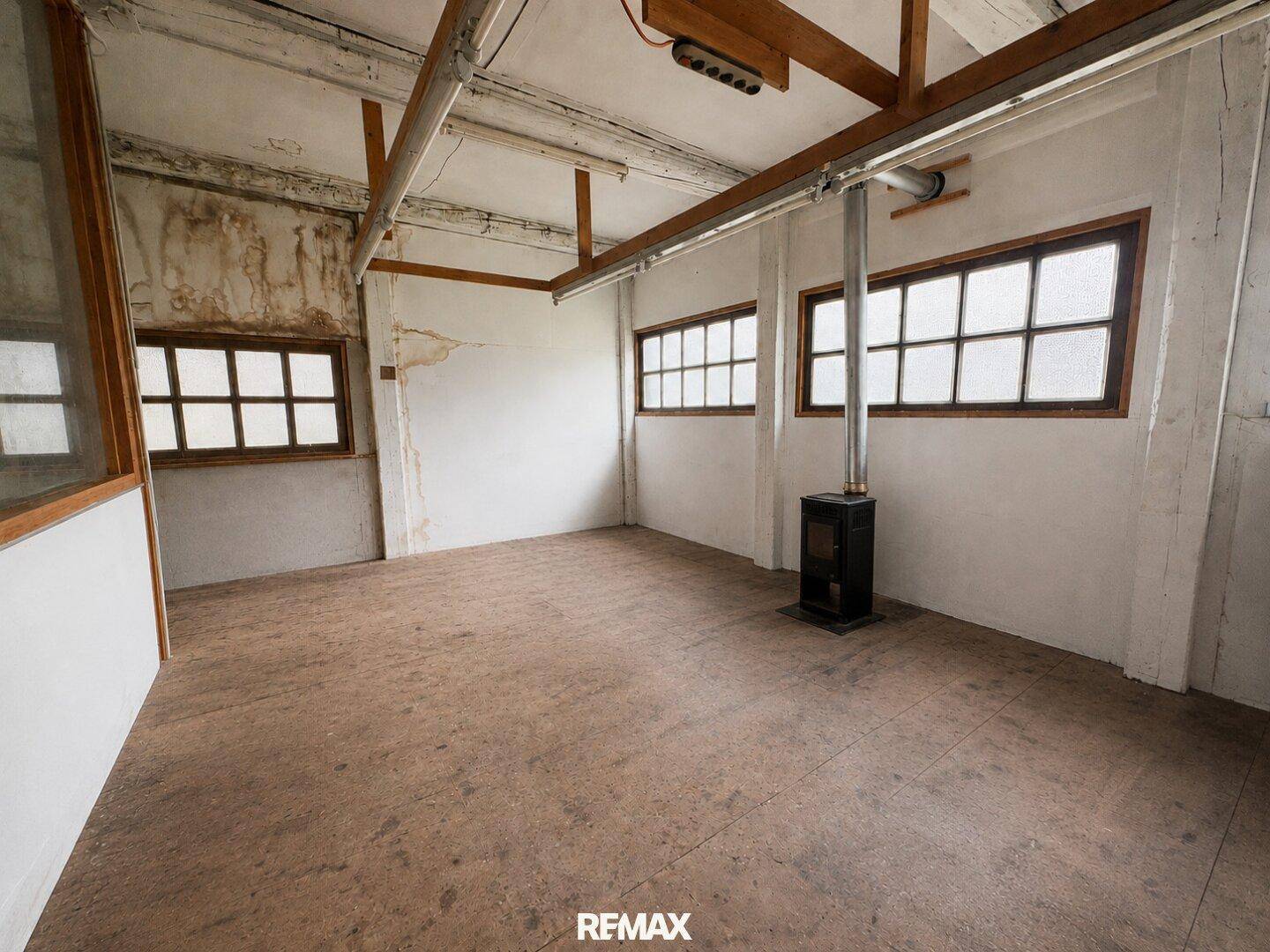 Heller Raum mit Holzofen, großen Fenstern und einfacher Ausstattung, ideal als Werkstatt.