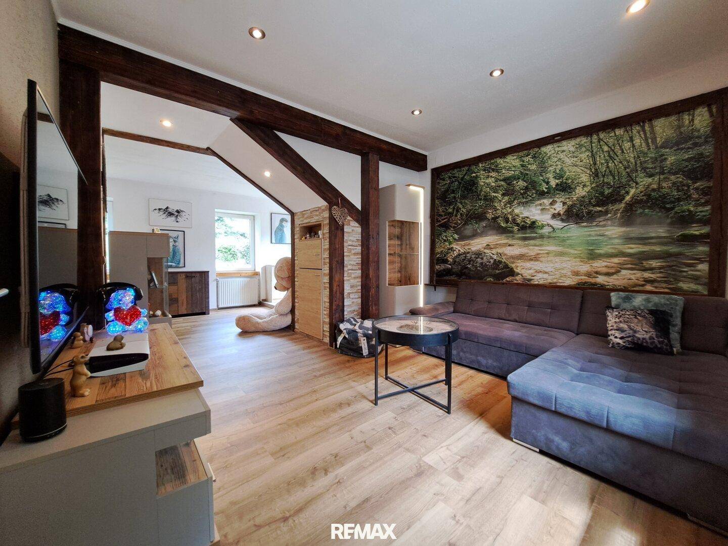Gemütliches Wohnzimmer mit Holzbalken, großem Sofa und einem Wandbild mit Naturmotiv.