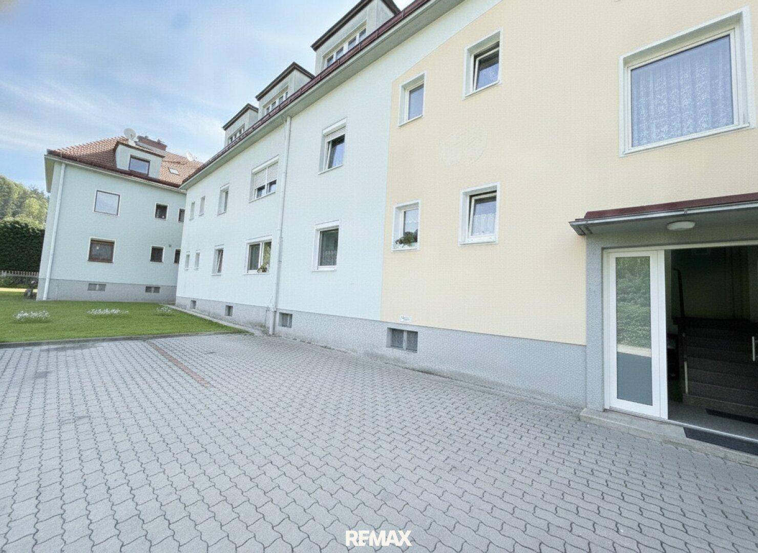 Mehrfamilienhaus mit heller Fassade, gepflastertem Parkplatz und Grünfläche.