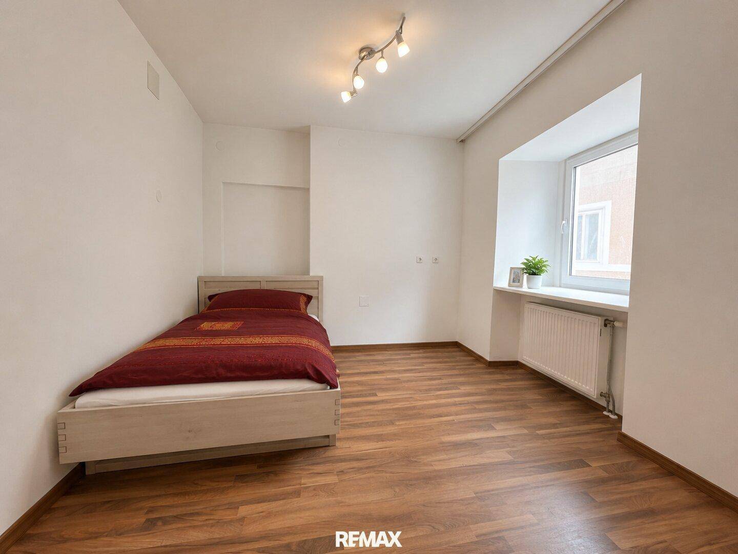 Helles Schlafzimmer mit Einzelbett, weißen Wänden und Holzoptik-Laminatboden.