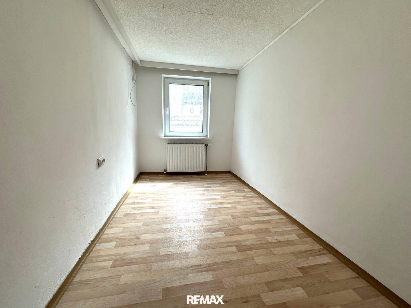 Leeres Zimmer mit hellem Laminatboden und einem Fenster, das Tageslicht hereinlässt.