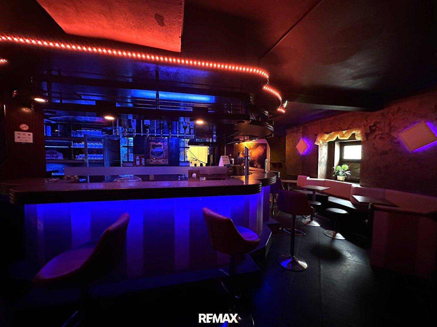 Bar-Bereich mit Neonbeleuchtung, Theke und Sitzgelegenheiten in dunkler Atmosphäre.