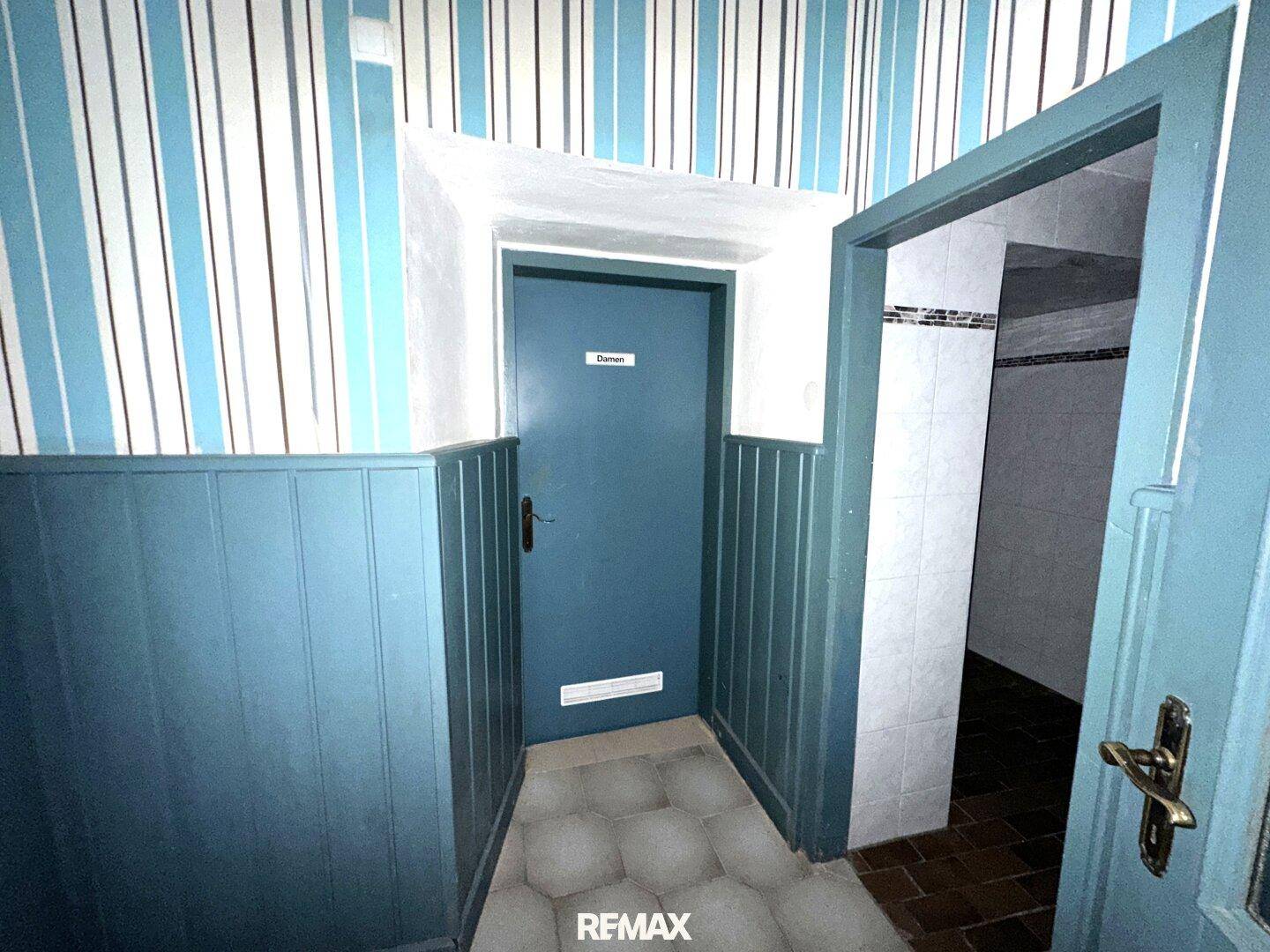 Eingangsbereich zu einem Damen-WC mit gestreifter Tapete und blauer Holzverkleidung in einem älteren Gebäude.