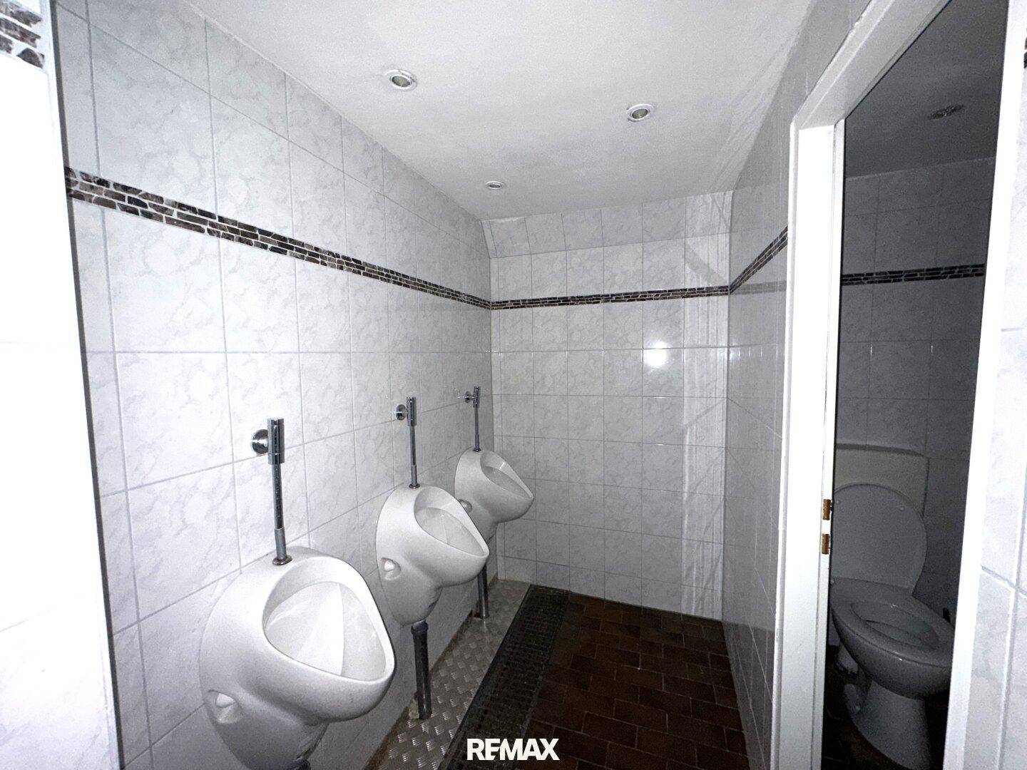 Gefliestes Herren-WC mit mehreren Urinalen und einer Toilette in einer separaten Kabine.