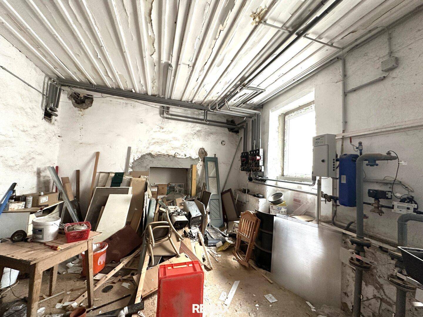 Unaufgeräumter Kellerraum mit sichtbaren Rohren, einem Fenster und technischer Ausstattung, die auf einen Hauswirtschaftsraum hindeutet.