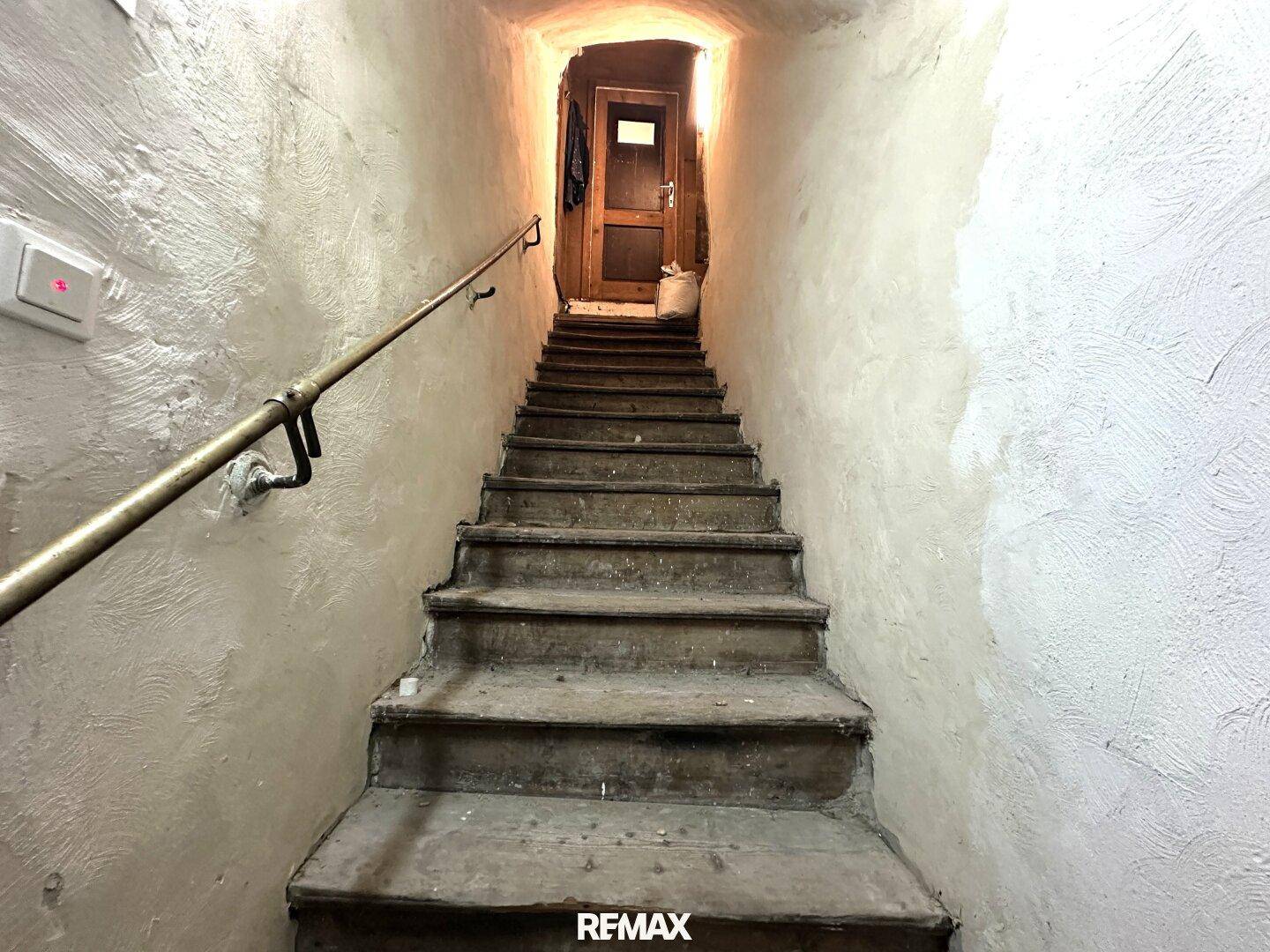 Alte, steile Holztreppe mit Handlauf, die in einen oberen, schwach beleuchteten Bereich führt.
