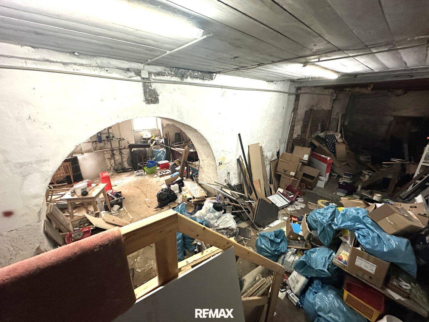 Großer, unaufgeräumter Kellerraum mit gewölbten Durchgängen und sichtbaren Holzdeckenbalken.