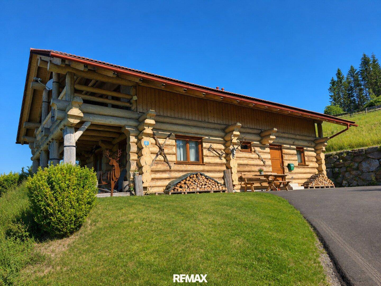 Blockhaus mit Holzlager und Sitzgelegenheit im Freien, umgeben von Grün und rustikalem Charme.