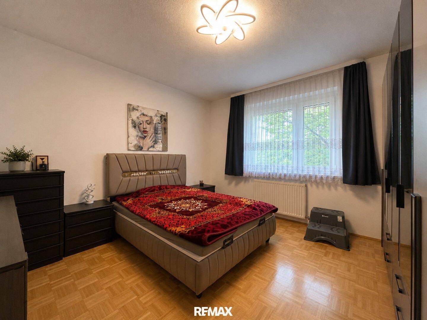 Helles Schlafzimmer mit Doppelbett, Parkettboden und großem Fenster mit Vorhängen.