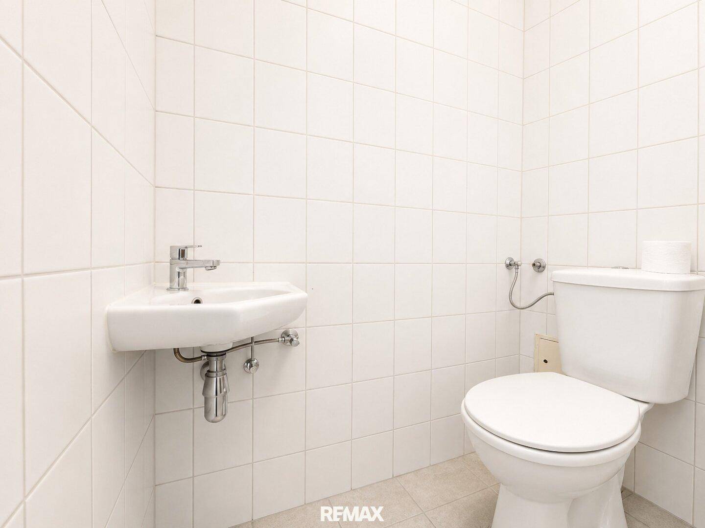 Kleines, weiß gefliestes Gäste-WC mit Toilette und Eckwaschbecken.